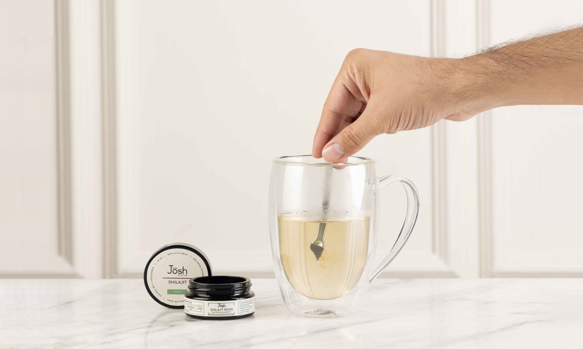 Hand mixing shilajit in a glassmug with hot water, next to a shilajit jar on a light surface.